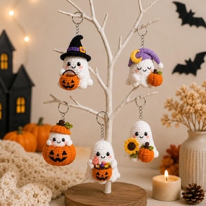 May include: Five white crochet ghost keychains hanging on a white decorative tree. One ghost wears a black witch hat, one a purple nightcap, one sits in a pumpkin, one holds a candy bucket, and one holds a sunflower and a small pumpkin.