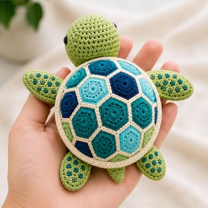 Op de afbeelding: Een kleine, handgemaakte gehaakte schildpad met een lichtgroene kop en ledematen met donkergroene accenten. Het schild is samengesteld uit meerdere zeshoekige motieven in diverse tinten blauw, groenblauw en donkerblauw, omlijnd met crèmekleurig garen.