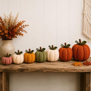May include: A row of seven handmade crochet pumpkins in varying sizes and colours, including dusty pink, cream, mustard yellow, sage green, white, and two shades of orange, each topped with a small brown stem and green crocheted leaves.