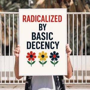 Op de afbeelding: Een rechthoekig bord met de tekst "RADICALIZED BY BASIC DECENCY" in rood en zwart. Daaronder drie gestileerde bloemen in rood, geel en blauw met groene stelen en bladeren. Het bord wordt door een persoon omhoog gehouden.