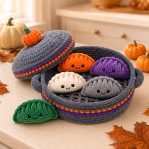 Puede incluir: Una vaporera de ganchillo gris con una tapa rematada por una pequeña calabaza naranja. Dentro y al lado de la cesta hay cinco pequeñas empanadillas de felpa en forma de media luna en naranja, morado, blanco, gris y verde.