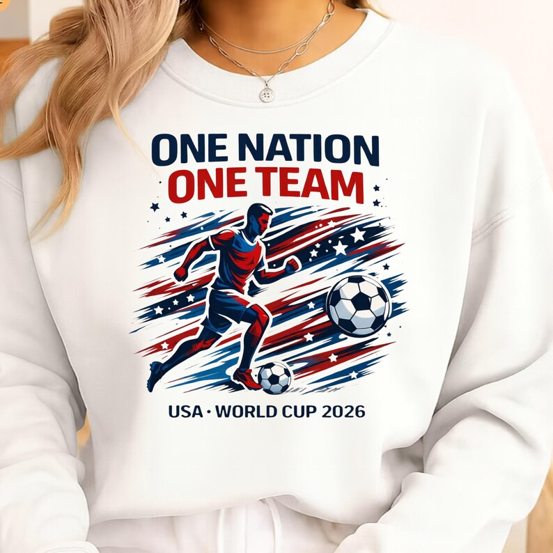 May include: White sweatshirt featuring a soccer player graphic in red and blue, kicking a soccer ball. Text reads "ONE NATION ONE TEAM" and "USA WORLD CUP 2026". Design incorporates stars and stripes.