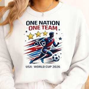 May include: White sweatshirt with a graphic of a soccer player in red, white, and blue, with the text "ONE NATION ONE TEAM" and "USA WORLD CUP 2026". The design includes stars and a soccer ball.