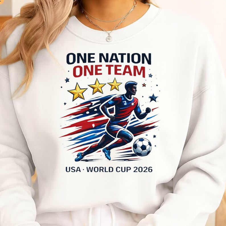 May include: White sweatshirt with a graphic of a soccer player in red, white, and blue, with the text "ONE NATION ONE TEAM" and "USA WORLD CUP 2026". The design includes stars and a soccer ball.