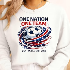 May include: White sweatshirt with a soccer ball graphic encircled by red, white, and blue swirls and stars. The text reads "ONE NATION ONE TEAM" and "USA WORLD CUP 2026".