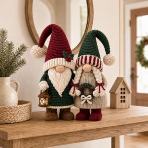 Puede incluir: Dos gnomos de ganchillo de pie uno al lado del otro. El gnomo de la izquierda lleva un gorro alto granate con un pompón, una túnica verde y sostiene un farol. El gnomo de la derecha tiene trenzas, un gorro a rayas verdes y granates y una corona.