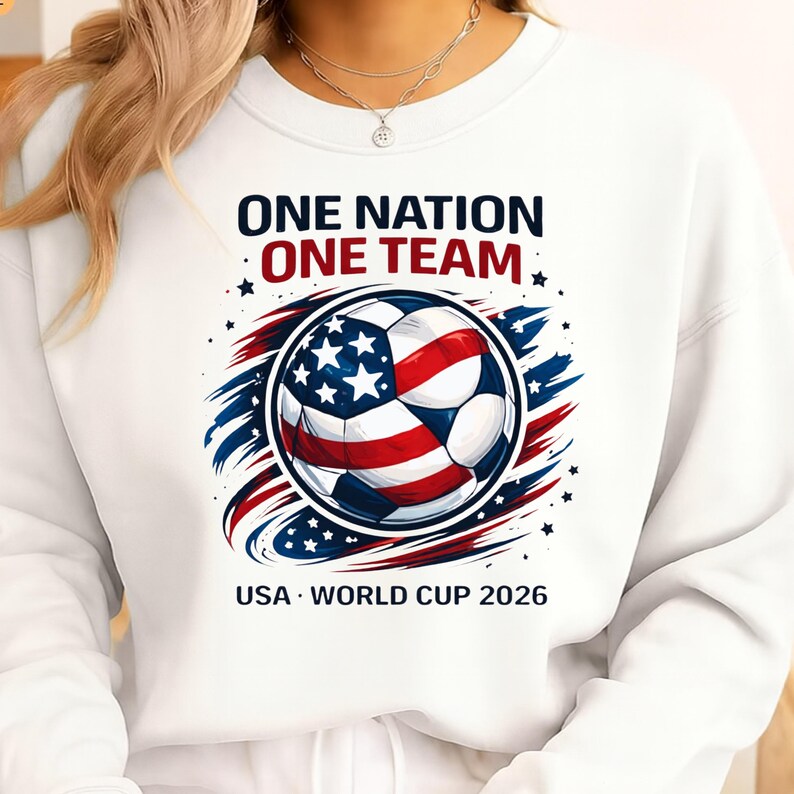 May include: White sweatshirt featuring a soccer ball design in red, white, and blue, with the American flag's stars and stripes. Text reads "ONE NATION ONE TEAM" and "USA WORLD CUP 2026".