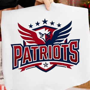 Pode incluir: Artigo de tecido branco com um design vermelho, branco e azul. O design inclui um emblema de águia, cinco estrelas e a palavra "PATRIOTS" em negrito. O tema geral sugere um artigo desportivo ou patriótico.