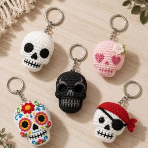 May include: Five crocheted skull keychains with metal rings. One is plain white with black details, one is pink with heart eyes and a flower, one is solid black, one features colourful floral embroidery, and one is a white skull wearing a red pirate bandana.