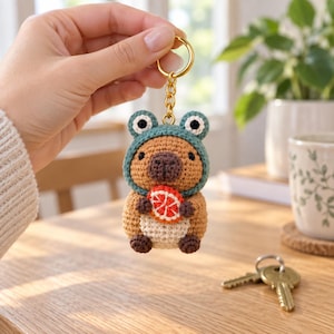 May include: A crocheted capybara keychain featuring a tan body with a cream-colored belly, dark brown paws, and a dark brown nose. The capybara wears a teal frog-shaped hood with white and black eyes and holds a small orange citrus slice.