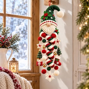 Könnte beinhalten: Eine hängende Häkel-Gnom-Dekoration mit einer grünen Zipfelmütze mit Stechpalmen, einem weißen Bart und roten Fäustlingen. Der Gnom ist mit baumelnden Ornamenten wie Zuckerstangen, Sternen, kleinen Weihnachtsbäumen, Geschenken und einer Schneeflocke verziert.