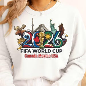May include: White sweatshirt featuring a colorful graphic for the 2026 FIFA World Cup. The design includes the number 2026, a trophy, and iconic landmarks from Canada, Mexico, and the USA. Text at the bottom reads "FIFA WORLD CUP Canada Mexico USA."