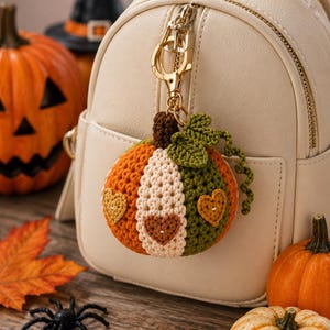 May include: A crocheted pumpkin keychain featuring orange, cream, and olive green segments with a brown stem and a green leaf. The pumpkin is decorated with small heart-shaped patches and is attached to a gold-toned metal lobster clasp.