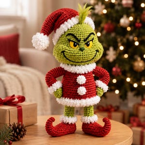 Könnte beinhalten: Eine grüne Häkelfigur mit gelben Augen und einem schelmischen Ausdruck, die eine rote Weihnachtsmütze mit weißem Rand, eine rote Jacke mit zwei weißen Bommeln und rote Schuhe mit aufgebogenen Spitzen und weißem Rand trägt.