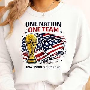 May include: White sweatshirt with a graphic featuring a gold trophy, an American flag heart, and the text "ONE NATION ONE TEAM" and "USA WORLD CUP 2026". The design is in red, white, blue, and gold.