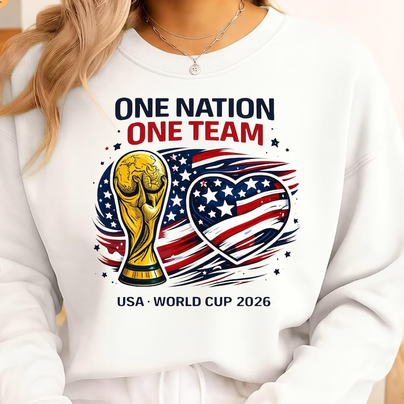 May include: White sweatshirt with a graphic featuring a gold trophy, an American flag heart, and the text "ONE NATION ONE TEAM" and "USA WORLD CUP 2026". The design is in red, white, blue, and gold.