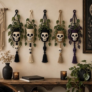 May include: Five wall-hanging crochet plant holders, each featuring a skull design on the pot and a smaller skull accent above a tassel. The holders contain crocheted green vines and are displayed in black, cream, black, tan, and purple color schemes.
