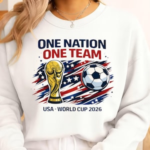 May include: White sweatshirt with the text "ONE NATION ONE TEAM" in navy and red. Features a gold trophy, soccer ball, and the text "USA - WORLD CUP 2026" in navy.