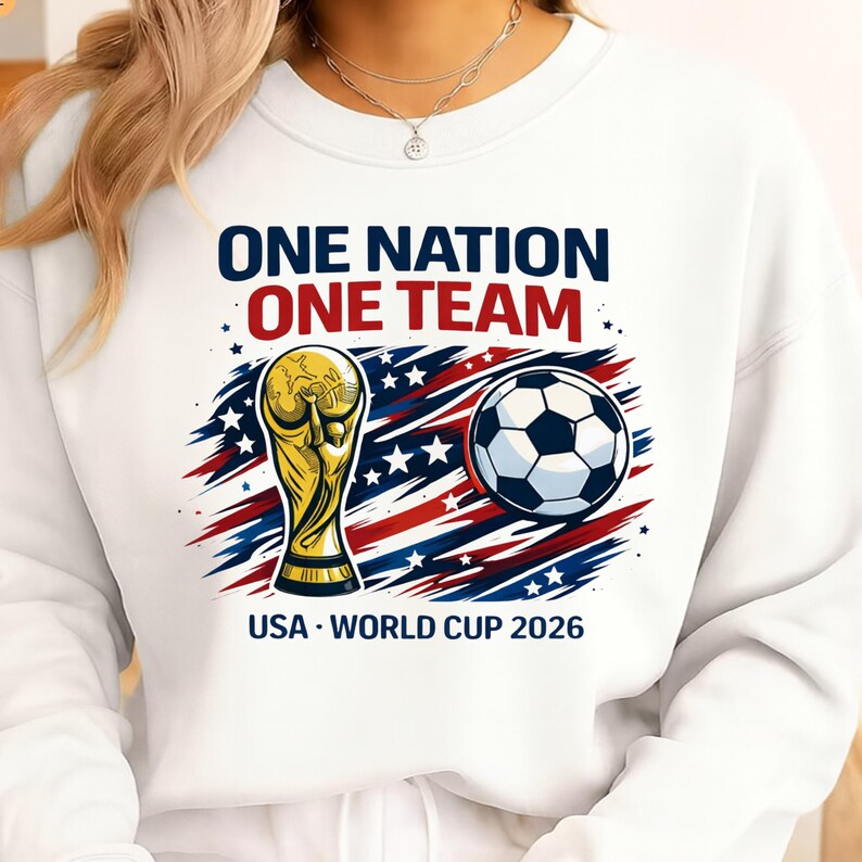 May include: White sweatshirt with the text "ONE NATION ONE TEAM" in navy and red. Features a gold trophy, soccer ball, and the text "USA - WORLD CUP 2026" in navy.