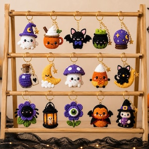 Könnte beinhalten: Ein hölzernes Präsentationsregal mit fünfzehn gehäkelten Schlüsselanhängern im Halloween-Design, darunter ein Geist, ein Kürbisbecher, eine Fledermaus, ein Kessel, eine Kristallkugel, ein Fläschchen, ein Halbmond, ein Pilz, ein Zuckermais, eine schwarze Katze, eine Spinne, eine Laterne, eine Blumenblüte mit Auge, ein Vampir und eine Hexe.