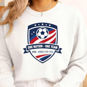 May include: White sweatshirt with a shield design. The design includes a soccer ball, stars, and the text "ONE NATION - ONE TEAM USA WORLD CUP 2026" in blue, red, and white. The design is centered on the front.