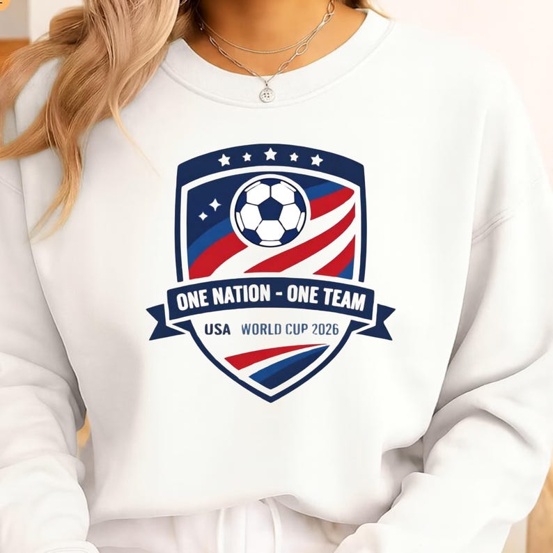 May include: White sweatshirt with a shield design. The design includes a soccer ball, stars, and the text "ONE NATION - ONE TEAM USA WORLD CUP 2026" in blue, red, and white. The design is centered on the front.