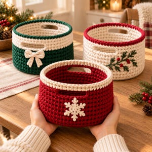 May include: Three round crochet baskets with integrated handles. One is dark green with a cream bow, one is cream with a green holly branch and red berries, and one is deep red with a cream snowflake appliqué, held by two hands.