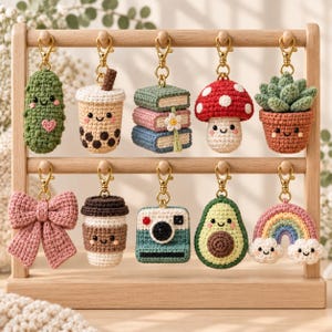 May include: A wooden display stand holding ten crocheted keychains featuring various designs including a pickle, boba tea, stacked books, a mushroom, a succulent, a pink bow, a coffee cup, a camera, an avocado, and a rainbow with clouds.