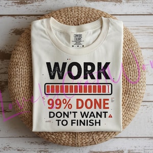 Könnte beinhalten: Cremefarbenes T-Shirt mit den Wörtern "WORK" und "99% DONE" in Schwarz bzw. Rot. Das Shirt zeigt eine Ladebalken-Grafik und den Satz "DON'T WANT TO FINISH". Das Shirt liegt gefaltet auf einer gewebten, kreisförmigen Oberfläche.