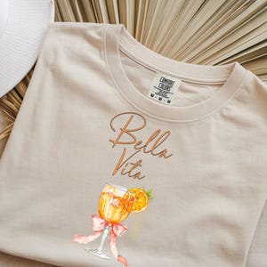 May include: Beige Comfort Colors t-shirt with the words "Bella Vita" in an elegant script. Below the text is a watercolor illustration of a cocktail with an orange slice and a pink bow. A white sun hat is visible in the upper left corner.