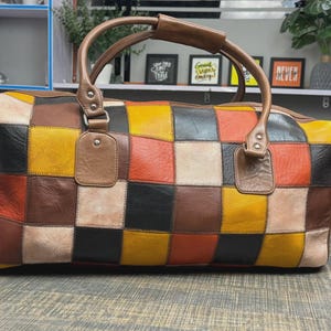 May include: A multi-coloured patchwork leather duffel bag with a brown leather handle. The bag features a design of squares in shades of brown, yellow, orange, and black. The bag has a zip closure and is designed for travel.