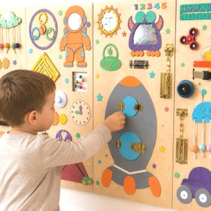 Puede incluir: Un gran tablero de actividades de madera con varios elementos interactivos, incluyendo un cohete con puertas que se abren, un reloj, engranajes, un espejo, un fidget spinner, pestillos de metal y formas coloridas como un astronauta y un monstruo.