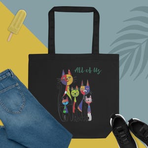 Może przedstawiać: Czarna torba typu tote z kolorową ilustracją sześciu stylizowanych kotów. Koty są w różnych kolorach, w tym czerwonym, żółtym, niebieskim i zielonym. Słowa "All of Us" są wydrukowane na zielono w prawym górnym rogu torby.