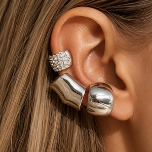 May include: A selection of silver ear cuffs and earrings. One cuff is set with small, sparkling stones. The other two earrings have a smooth, polished finish with a geometric design. The jewellery is worn on the ear.