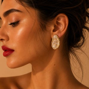May include: An irregular, oval-shaped stud earring featuring a smooth, pearlescent white centre with a marbled texture, encased in a textured, metallic gold-toned border. The earring is worn on the earlobe, showcasing its organic, asymmetrical form.