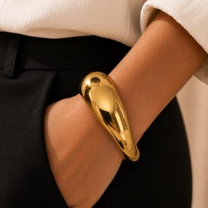May include: A polished gold-tone cuff bracelet with a smooth, rounded, and organic teardrop shape. The metal surface is highly reflective, creating a bright sheen that wraps around the wrist, showcasing a solid, continuous, and seamless design.