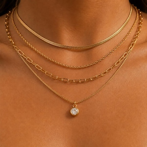 Puede incluir: Un conjunto de collares dorados en capas. Los collares incluyen una cadena de espiga, una cadena delicada, una cadena con eslabones rectangulares y un collar con colgante con una piedra preciosa redonda y transparente. Los collares se muestran en un cuello.