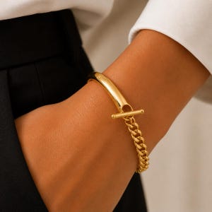 May include: A gold-toned bracelet featuring a smooth, curved solid bar section connected to a polished curb chain. The two segments are joined by a horizontal toggle bar clasp mechanism that secures the chain to the solid metal component.