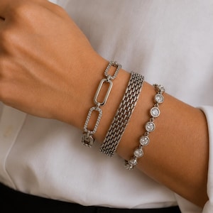 May include: A set of three silver-toned bracelets. One features rectangular links with embedded clear stones, the second is a wide woven mesh band, and the third is a delicate chain with a series of round, bezel-set clear stones.