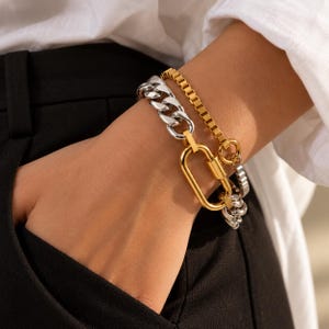 May include: A two-tone bracelet set featuring a thick silver-coloured curb chain and a thinner gold-coloured box chain. The two chains are connected by a large, oval-shaped gold-coloured carabiner-style clasp that serves as a central focal point.