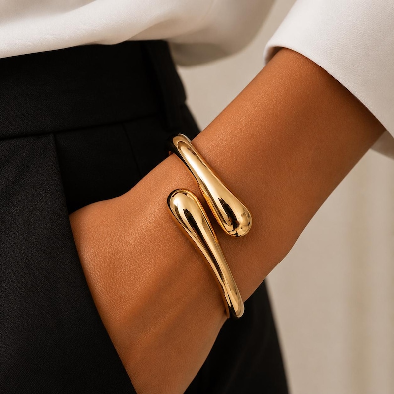 May include: A polished gold-tone cuff bracelet featuring a bypass design with two smooth, rounded, teardrop-shaped ends that overlap slightly. The metal surface is reflective and solid, creating a continuous, fluid, and minimalist circular form.