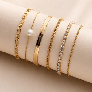 Può includere: Una collezione di braccialetti color oro esposti su una superficie neutra. I braccialetti presentano vari design, tra cui una catena a maglie, una catena delicata con una perla, un motivo a spina di pesce, una torsione a corda, una fila di pietre trasparenti e una catena sottile.