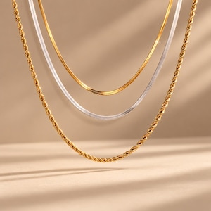 May include: A layered necklace set consisting of three distinct chains. The outermost is a thick gold-toned rope chain, the middle is a silver-toned flat herringbone chain, and the innermost is a thin gold-toned flat herringbone chain.