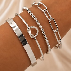 Può includere: Una collezione di quattro braccialetti d'argento. Uno è una fascia larga e piatta, un altro è una fascia sottile con un design a nodo, un terzo è un braccialetto di perline e l'ultimo è un braccialetto a catena. I braccialetti sono indossati su un braccio.