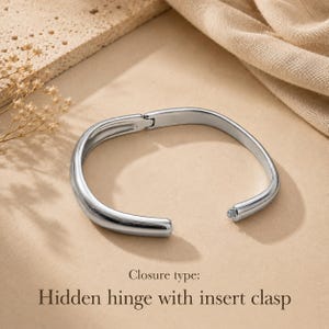 May include: A polished silver-tone metal bangle bracelet with a smooth, rounded profile. The bracelet features a functional hidden hinge mechanism on one side and an insert clasp closure on the other, as indicated by the text Closure type: Hidden hinge with insert clasp.