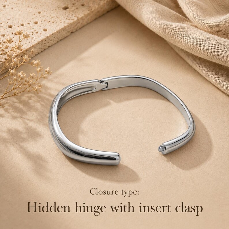 May include: A polished silver-tone metal bangle bracelet with a smooth, rounded profile. The bracelet features a functional hidden hinge mechanism on one side and an insert clasp closure on the other, as indicated by the text Closure type: Hidden hinge with insert clasp.