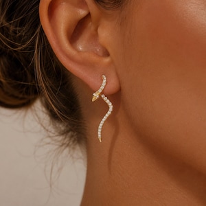 May include: A gold-toned snake-shaped earring featuring a curved body encrusted with small, clear, sparkling stones. The earring is designed to follow the natural contour of the earlobe, with the head of the snake pointing upwards.