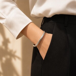 May include: A smooth, polished silver-tone bangle bracelet with a rounded, tubular profile. The bracelet is worn on the wrist, positioned near the pocket of black trousers and a white long-sleeved shirt, showcasing a clean, reflective metallic finish.