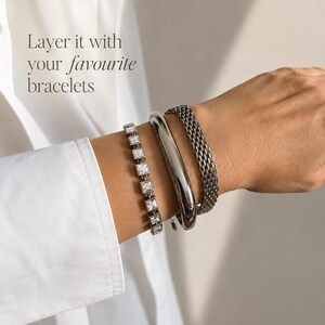 May include: Three silver-toned bracelets worn together: one featuring square-cut clear stones, one smooth polished bangle, and one textured mesh chain. The text "Layer it with your favourite bracelets" is visible in the upper left corner.
