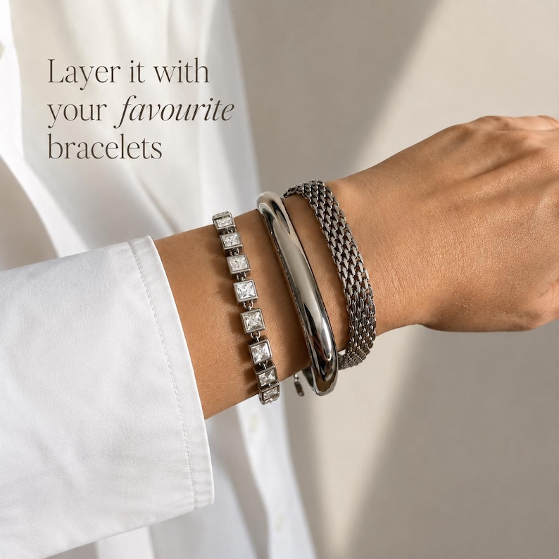 May include: Three silver-toned bracelets worn together: one featuring square-cut clear stones, one smooth polished bangle, and one textured mesh chain. The text "Layer it with your favourite bracelets" is visible in the upper left corner.
